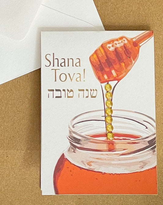 this is a mini note card on a craft paper gift bag with a white envelope and a black marker , the card says Shana Tova in English and Hebrew with a glass jar of honey and honey pouring down from the honey server