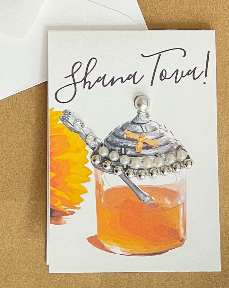 this is a mini note card on a craft paper gift bag with a white envelope and a black marker , the card says Shana Tova in English with a glass jar of honey 